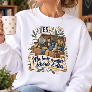 May include: A white long-sleeve sweatshirt featuring a graphic of a wooden box filled with a magnifying glass, hourglass, and various shapes. The text reads "TES" at the top and "Ma boîte à outils déborde d'idées" on a banner at the bottom, surrounded by floral accents.