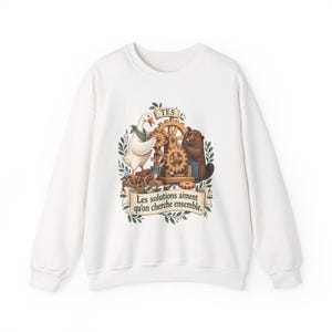 May include: A white long-sleeve sweatshirt featuring a central graphic of a goose, a mouse, and a beaver working together on a wooden gear mechanism. The text "TES" appears above the gears, and a banner below reads "Les solutions aiment qu'on cherche ensemble."