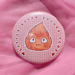 May include: A round button with the text "LITTLE BACON BURNER CLUB" encircling a cartoon flame character. The button has a red and white plaid background and is set against a pink fabric.