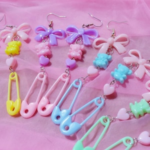 Könnte beinhalten: Ein Set mit sechs Paaren bunter Hängeohrringe mit Sicherheitsnadel-Anhängern, Schleifen, Herzen und Bärenanhängern. Die Ohrringe sind aus Kunststoff und in Pink, Gelb, Blau und Grün erhältlich.