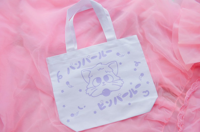 Puede incluir: Bolsa de tela blanca con un dise&ntilde;o de gato de dibujos animados morado y el texto "パジャマパロール" en japon&eacute;s. La bolsa tiene un asa blanca.