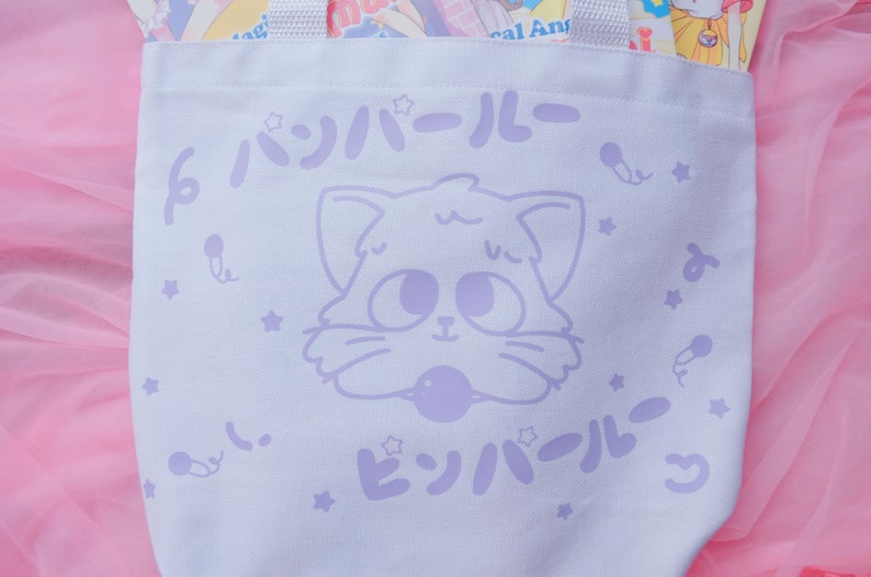 Puede incluir: Bolsa de lona blanca con un dise&ntilde;o de gato de dibujos animados morado y el texto "ビッハイル" en caracteres japoneses. La bolsa tiene un patr&oacute;n de estrellas moradas.