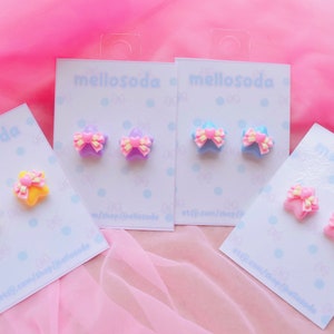 Könnte beinhalten: Vier Paare sternförmiger Ohrringe mit Schleifen. Die Ohrringe sind in Pink, Blau und Gelb mit rosa Schleifen. Die Ohrringe werden auf weißen Karten mit dem Text "mellosoda" darauf präsentiert.