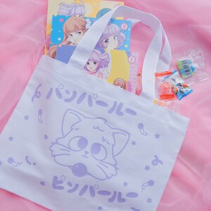 Puede incluir: Una bolsa de tela blanca con una ilustraci&oacute;n de un gato de dibujos animados y el texto "ピンボール" en japon&eacute;s. La bolsa tiene asas blancas y est&aacute; sobre un fondo rosa.