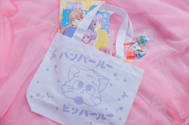 Puede incluir: Una bolsa de tela blanca con una ilustraci&oacute;n de un gato de dibujos animados y el texto "ピンボール" en japon&eacute;s. La bolsa tiene asas blancas y est&aacute; sobre un fondo rosa.