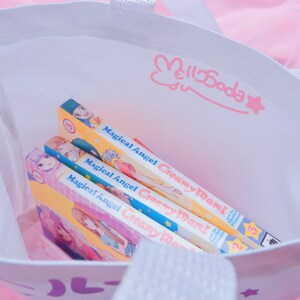 Puede incluir: Una bolsa de tela blanca con un dise&ntilde;o de conejo rosa y estrella. La bolsa est&aacute; llena de tres vol&uacute;menes de manga de "Magical Angel Creamy Mami" con portadas amarillas, rosas y azules.