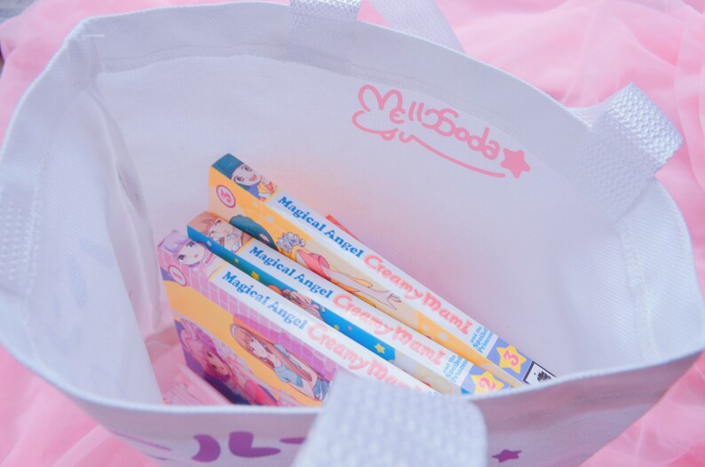 Puede incluir: Una bolsa de tela blanca con un dise&ntilde;o de conejo rosa y estrella. La bolsa est&aacute; llena de tres vol&uacute;menes de manga de "Magical Angel Creamy Mami" con portadas amarillas, rosas y azules.