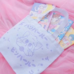 Puede incluir: Una bolsa de tela blanca con un gato de dibujos animados morado y texto japon&eacute;s que dice "Twin-Tail". La bolsa tiene un asa blanca y est&aacute; llena de libros de manga.