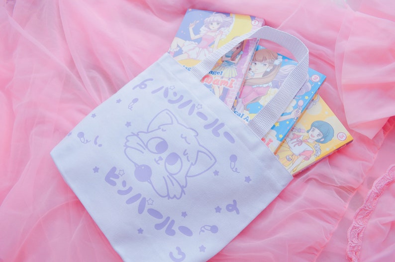 Puede incluir: Una bolsa de tela blanca con un gato de dibujos animados morado y texto japon&eacute;s que dice "Twin-Tail". La bolsa tiene un asa blanca y est&aacute; llena de libros de manga.