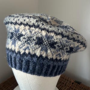 Puede incluir: Boina de punto en tonos azules, crema y gris, con un diseño estampado. El borde es de color azul oscuro liso y el sombrero se exhibe sobre una cabeza de maniquí blanca. La boina está hecha de un material suave y texturizado.