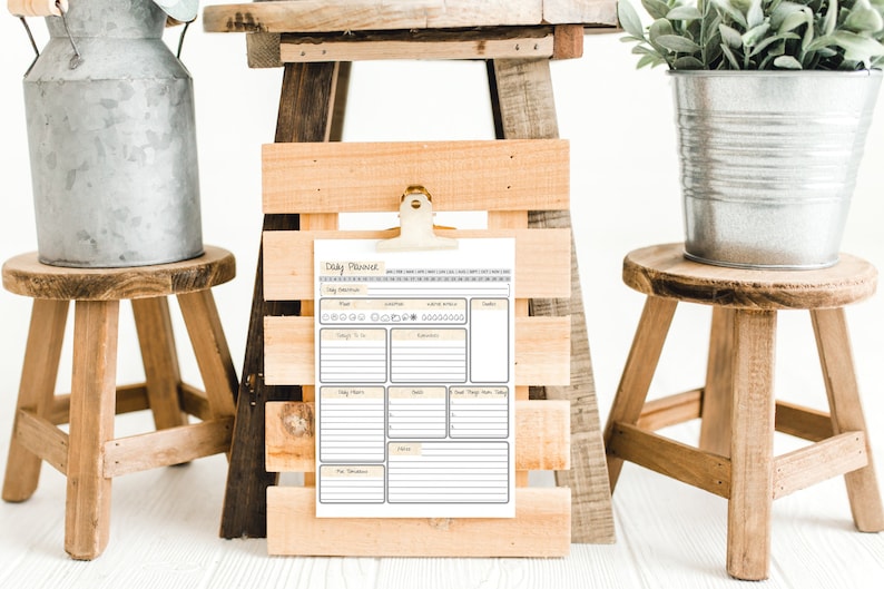 May include: A daily planner with a yellow and brown colour scheme is clipped to a wooden frame. The planner has sections for daily tasks, meal planning, and notes. The frame is sitting on a wooden stool.