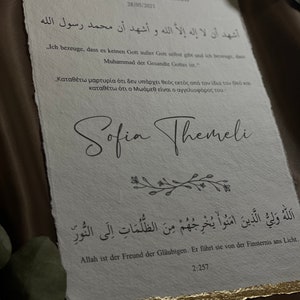 May include: A white certificate with gold accents, featuring Arabic text and a signature. The text reads "Ash-hadu alla ilaha illa-llah wa ash-hadu anna Muhammadan rasul-Allah."  The certificate is titled "Zertifikat zur Konversion" and dated 28/05/2021. The signature reads "Sofia Themeli".