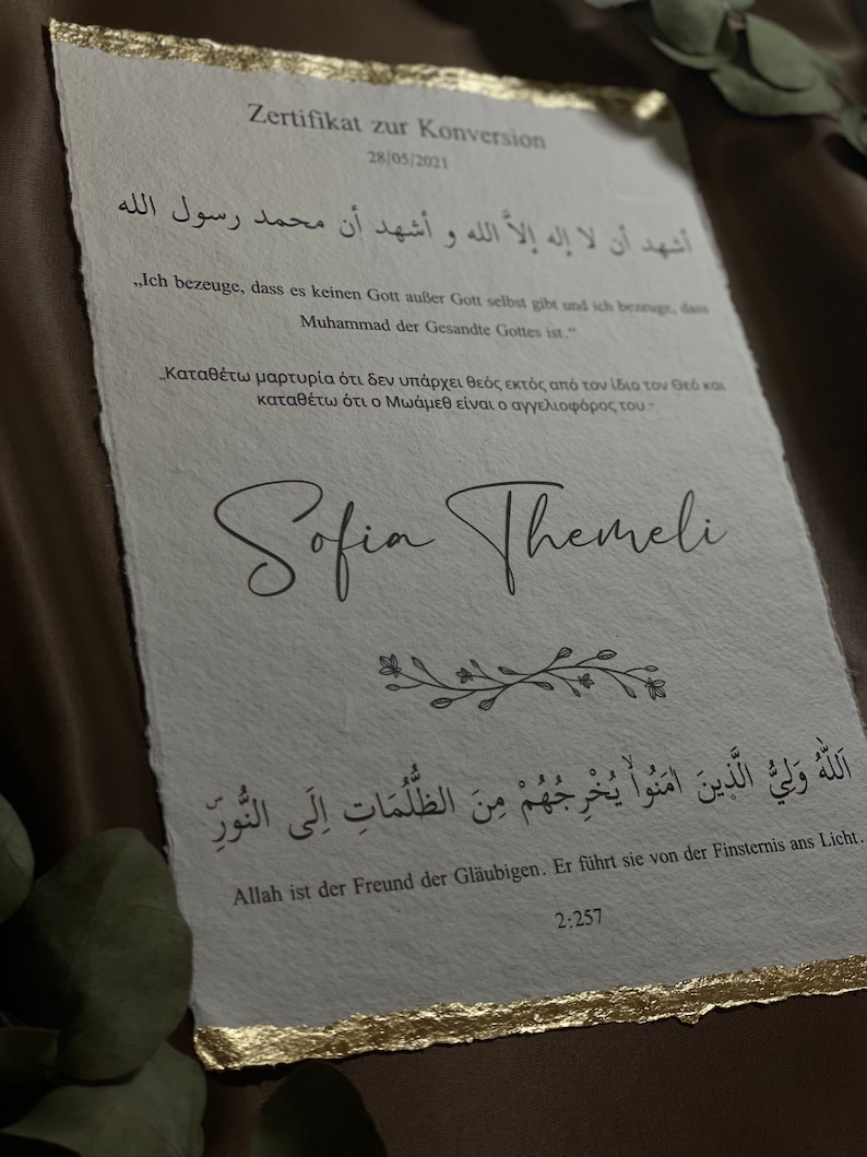 May include: A white certificate with gold accents, featuring Arabic text and a signature. The text reads "Ash-hadu alla ilaha illa-llah wa ash-hadu anna Muhammadan rasul-Allah."  The certificate is titled "Zertifikat zur Konversion" and dated 28/05/2021. The signature reads "Sofia Themeli".