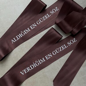 Pode incluir: Duas fitas de cetim castanho-escuro com texto impresso em branco. Uma fita exibe a frase ALDIĞIM EN G&Uuml;ZEL S&Ouml;Z, enquanto a outra fita exibe a frase VERDİĞİM EN G&Uuml;ZEL S&Ouml;Z. As fitas t&ecirc;m um acabamento liso e reflexivo e est&atilde;o dispostas.