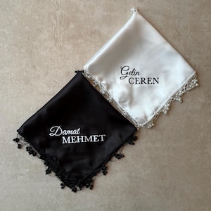 Puede incluir: Se muestran dos pañuelos de satén, uno negro y otro blanco. El pañuelo negro tiene el nombre "Damat MEHMET" bordado en blanco, mientras que el blanco tiene "Gelin CEREN" en negro. Ambos están adornados con encaje de cuentas.