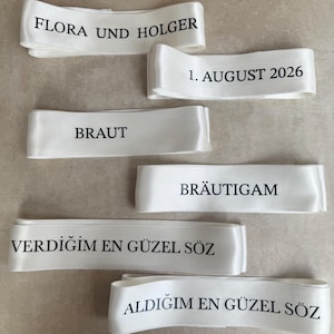 Pode incluir: Seis fitas de cetim branco com texto impresso em preto. As fitas exibem as frases: FLORA UND HOLGER, 1. AUGUST 2026, BRAUT, BR&Auml;UTIGAM, VERDIĞIM EN G&Uuml;ZEL S&Ouml;Z e ALDIĞIM EN G&Uuml;ZEL S&Ouml;Z. Cada fita tem um acabamento liso e reflexivo.