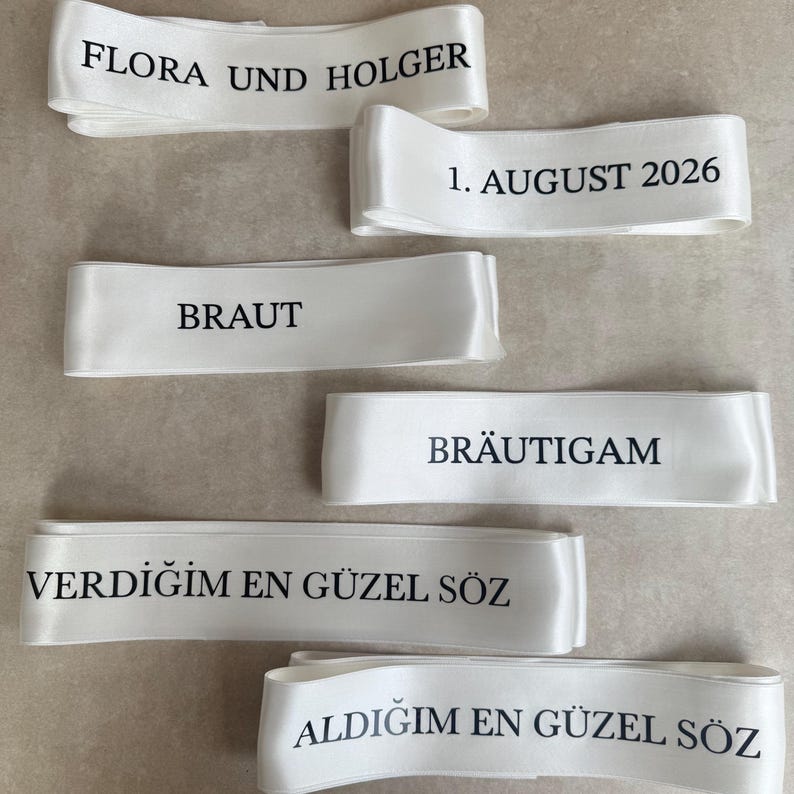 Pode incluir: Seis fitas de cetim branco com texto impresso em preto. As fitas exibem as frases: FLORA UND HOLGER, 1. AUGUST 2026, BRAUT, BR&Auml;UTIGAM, VERDIĞIM EN G&Uuml;ZEL S&Ouml;Z e ALDIĞIM EN G&Uuml;ZEL S&Ouml;Z. Cada fita tem um acabamento liso e reflexivo.
