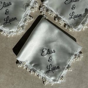 Puede incluir: Tres pañuelos de satén blanco con ribete de perlas y los nombres "Elisa & Luan" bordados en cada uno.