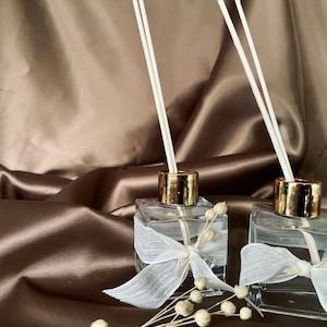 Könnte beinhalten: Zwei transparente Glas-Duftzerstäuber mit goldfarbenen Kragen und weißen Stäbchen. Jede Flasche ist mit einem weißen Band verziert. Getrocknete weiße Blumen sind in der Nähe der Flaschen angeordnet, vor einem braunen Satinhintergrund. Ein Raumduftprodukt.