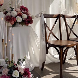 Pode incluir: Uma cadeira de madeira castanho-escura com encosto em cruz, decorada com uma fita de cetim branca atada na lateral com um la&ccedil;o. A fita tem impresso o texto preto VERDİĞİM EN G&Uuml;ZEL S&Ouml;Z. A cadeira est&aacute; posicionada junto a uma mesa com tecido branco e flores.
