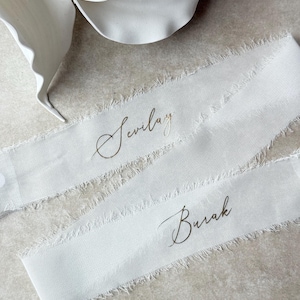 May include: Two white ribbons with frayed edges, each bearing a name in elegant gold script: "Sevilay" and "Burak." The ribbons are arranged on a neutral-toned surface, with a white flower in the background.
