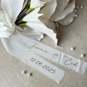 May include: A white floral arrangement with a satin ribbon and scattered pearls. The ribbon is inscribed with names and a date in gold lettering. The flowers have large, layered petals and are tied with a bow.