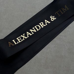 Pode incluir: Uma fita de cetim preta com o texto ALEXANDRA & TIM impresso numa fonte dourada met&aacute;lica. A fita est&aacute; posicionada na diagonal no enquadramento, exibindo uma textura lisa e reflexiva e uma tipografia clara e em negrito para os nomes.