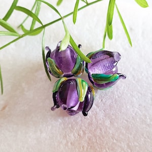 May include: Three purple glass beads with green and white accents, shaped like flowers. The beads are on a white surface.