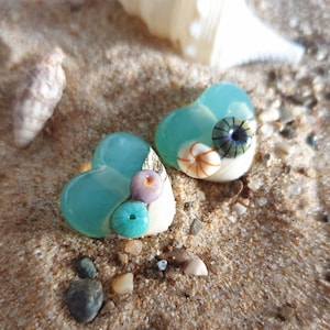 May include: Two heart-shaped resin pendants with a blue-green color. Each pendant is decorated with small seashells and a blue and white swirl design.