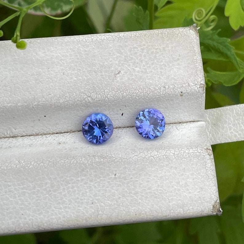 Puede incluir: Dos gemas azules redondas y facetadas se muestran en una caja blanca texturizada. Las gemas tienen un color azul profundo y vibrante con facetas internas visibles. El fondo es de un verde suave.