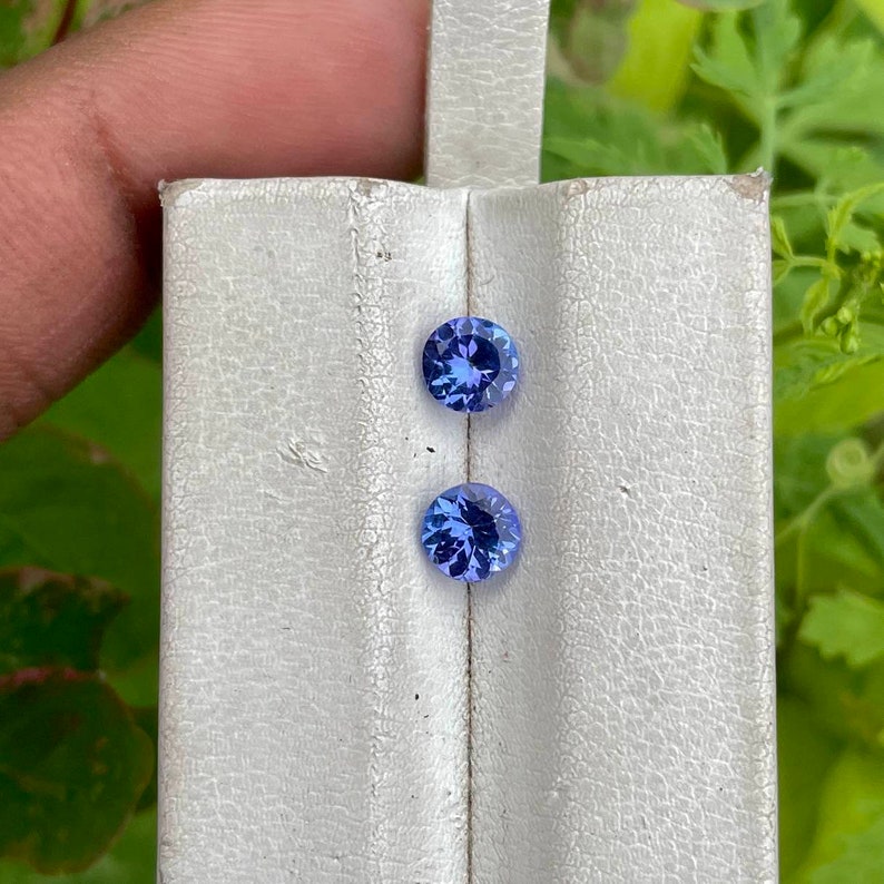 Puede incluir: Dos piedras preciosas azules redondas y facetadas se muestran sobre una superficie blanca texturizada. Las gemas son de un tono azul vibrante, con facetas visibles que capturan la luz. El fondo est&aacute; borroso, con follaje verde.