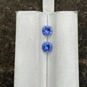 Puede incluir: Dos gemas azules redondas y facetadas se muestran sobre una superficie blanca texturizada. Las gemas tienen un corte brillante, mostrando su estructura interna y color. Las piedras preciosas son probablemente para la fabricaci&oacute;n de joyas.