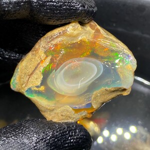 Könnte beinhalten: Ein roher Opal-Edelstein mit einem lebendigen Farbspiel. Der Opal hat einen milchig-weißen Kern, der von einer durchscheinenden, irisierenden Schicht umgeben ist. Die äußeren Ränder sind eine raue, hellbraune Matrix.