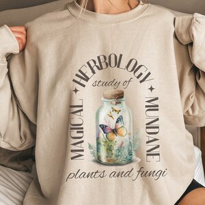 May include: A beige sweatshirt with a graphic design of a glass jar with butterflies and plants. The text around the jar reads "Herbology, study of magical and mundane plants and fungi."
