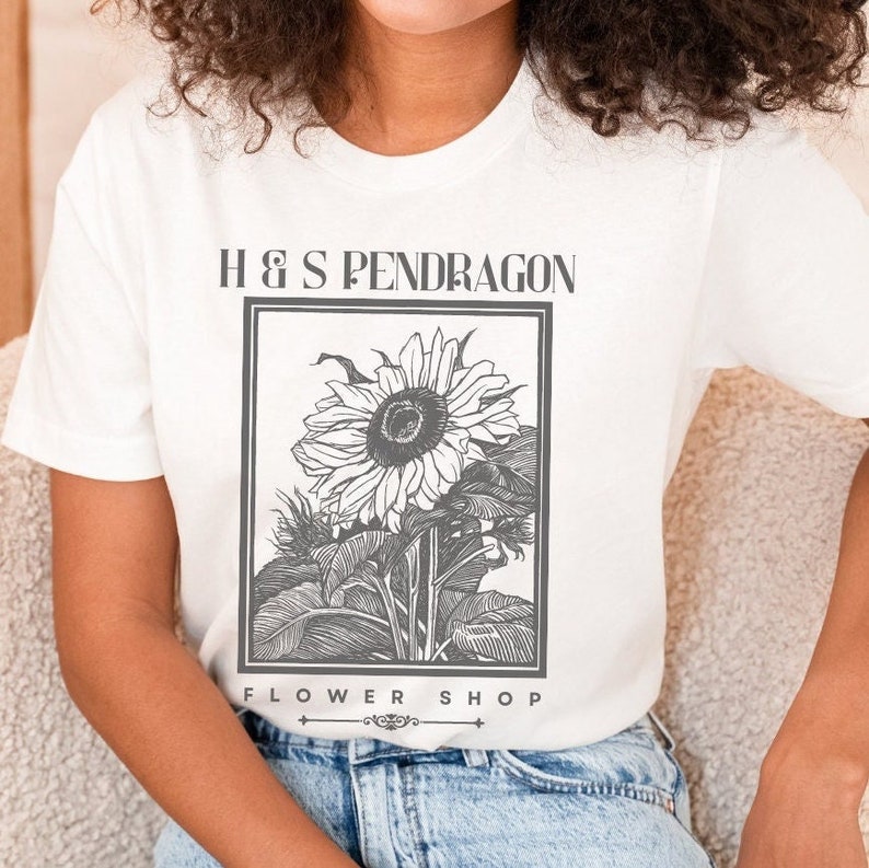 Peut inclure: T-shirt blanc avec un graphique noir et blanc d'un tournesol et le texte "H & S Pendragon Flower Shop".