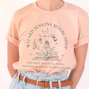 May include: A peach-colored t-shirt with a black graphic design of a book, flowers, a crescent moon, and an hourglass. The text "Wizard Jenkins Magic Shop" is written in a circle around the graphic. The text "The best magical items, potions & spells in Porthaven" is written below the graphic.