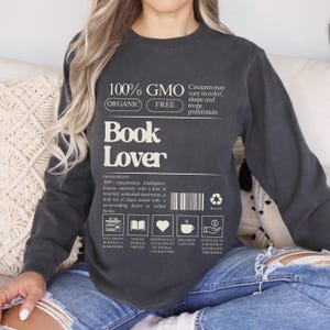 May include: Dark gray long-sleeve shirt with the text "Book Lover" in white. The shirt also features the text "100% GMO Free" and a list of ingredients, including imagination and intelligence. The shirt has a recycling symbol and icons related to books.
