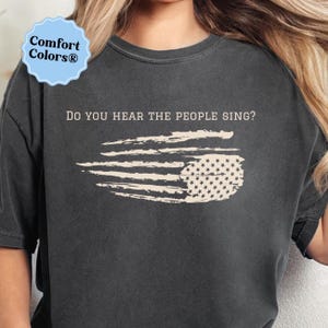May include: Dark gray t-shirt with the text "DO YOU HEAR THE PEOPLE SING?" and a distressed American flag design. The shirt features a round neck and short sleeves. The Comfort Colors® logo is visible in the upper left corner.