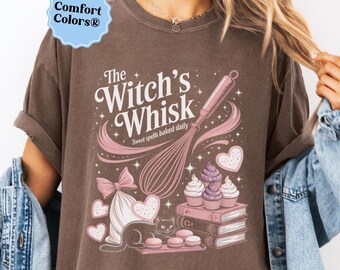 May include: Brown t-shirt with a graphic featuring a whisk, cupcakes, and the text "The Witch's Whisk Sweet spells baked daily." The design includes a cat, hearts, and a bow. The shirt has a Comfort Colors® label.