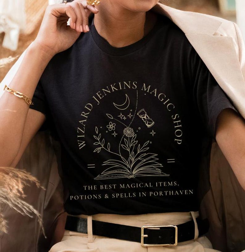 May include: Black t-shirt with a gold graphic design that says "Wizard Jenkins Magic Shop" with a crescent moon, stars, a book, and an hourglass. The text below the graphic says "The best magical items, potions & spells in Porthaven."