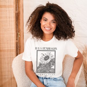Peut inclure: Un t-shirt blanc avec une illustration en noir et blanc d'un tournesol. Le texte "H & S PENDRAGON FLOWER SHOP" est imprim&eacute; sur le t-shirt.