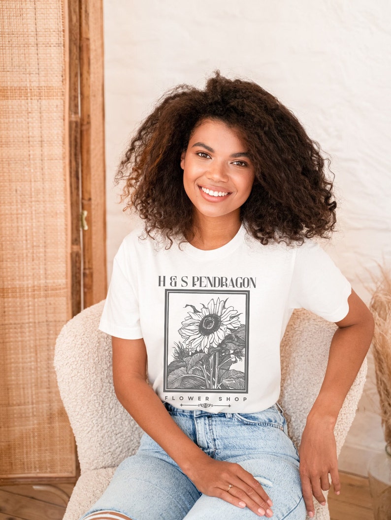 Peut inclure: Un t-shirt blanc avec une illustration en noir et blanc d'un tournesol. Le texte "H & S PENDRAGON FLOWER SHOP" est imprim&eacute; sur le t-shirt.