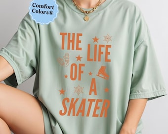 May include: Sage green t-shirt with the text "THE LIFE OF A SKATER" in orange. The design includes stars, a mitten, a snowflake, and a figure skate. The shirt has a relaxed fit and short sleeves.
