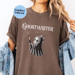 May include: Brown t-shirt with the word "Ghostwriter" in white script. A cartoon ghost holding a pen is featured below the text. The ghost is wearing a decorative shawl and bow. The shirt has the "Comfort Colors" logo.