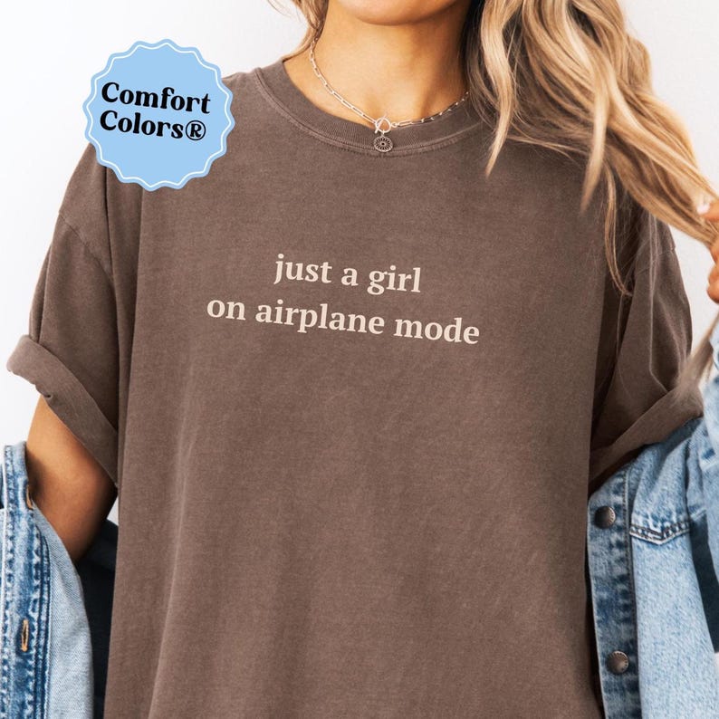Puede incluir: Una camiseta marr&oacute;n Comfort Colors con el texto "just a girl on airplane mode" en una fuente sencilla. La camiseta tiene las mangas enrolladas y un corte relajado. Una chaqueta vaquera es visible en el lateral.