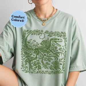 May include: Sage green t-shirt featuring a green dinosaur skeleton design surrounded by floral elements. The design is centered on the shirt. The shirt has short sleeves and a crew neck. The Comfort Colors logo is visible in the upper left corner.