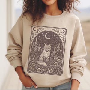 May include: A beige crewneck sweatshirt featuring a dark brown rectangular graphic print. The design depicts a fox sitting in a forest surrounded by flowers, pine trees, stars, and a crescent moon within an arched frame with leafy borders.