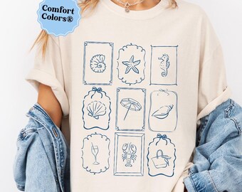 May include: Cream-colored Comfort Colors t-shirt featuring nine blue framed illustrations of seashells, a starfish, a seahorse, an umbrella, a champagne flute, a lobster, and a beach bag. The shirt has the Comfort Colors logo in the upper left corner.