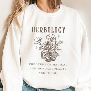 May include: White sweatshirt with the text "HERBOLOGY" in brown lettering. A black and white illustration of a book with flowers growing out of it. The text "THE STUDY OF MAGICAL AND MUNDANE PLANTS AND FUNGI." is below the illustration.