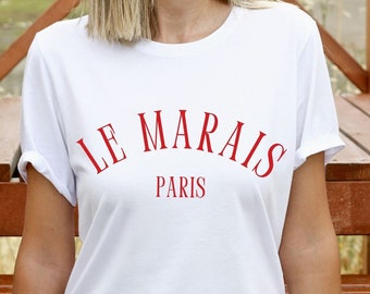 May include: White t-shirt with red text that reads "LE MARAIS PARIS". The person wearing the shirt is sitting on wooden steps.